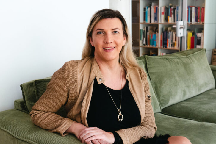 Julia Monro sitzt auf einem grünen Sofa und lächelt leicht in die Kamera. Im Hintergrund sind Bücherregale.