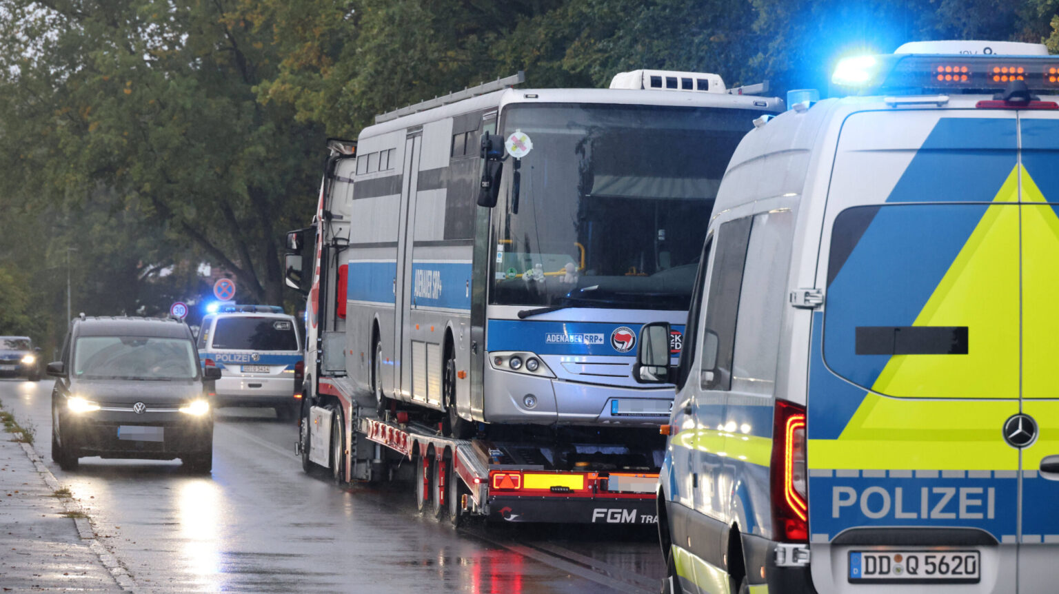 Polizeifahrzeuge mit Blaulicht auf nasser Straße, darunter ein großer Gefangenentransporter auf Anhänger, umgeben von Bäumen.