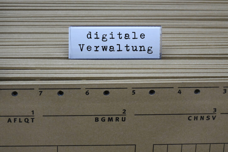 Auf Rücken von gelben Aktenordnern ist ein Trenner zu sehen. Auf ihm steht "digitale Verwaltung" geschrieben.