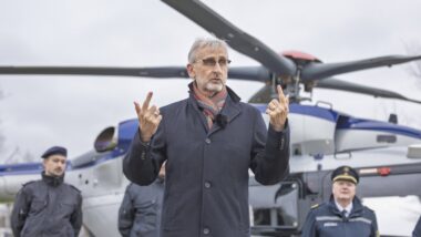 Ein Mann in einem blauen Mantel steht vor einem Helikopter und hält an beiden Händen Daumen und Zeigefinger in die Höhe.