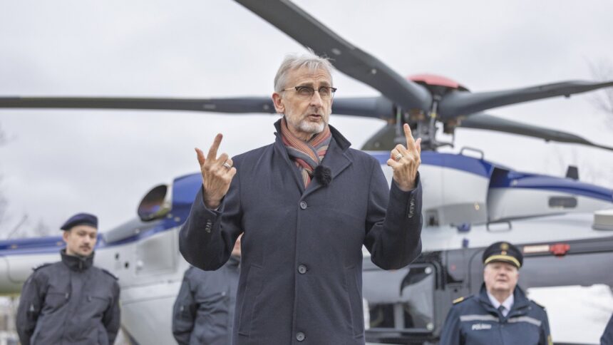 Ein Mann in einem blauen Mantel steht vor einem Helikopter und hält an beiden Händen Daumen und Zeigefinger in die Höhe.