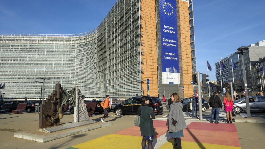 Bild des Gebäudes der EU-Kommission. Menschen stehen auf einem regenbogenfarbenden Fußgängerüberweg vor dem Gebäude.