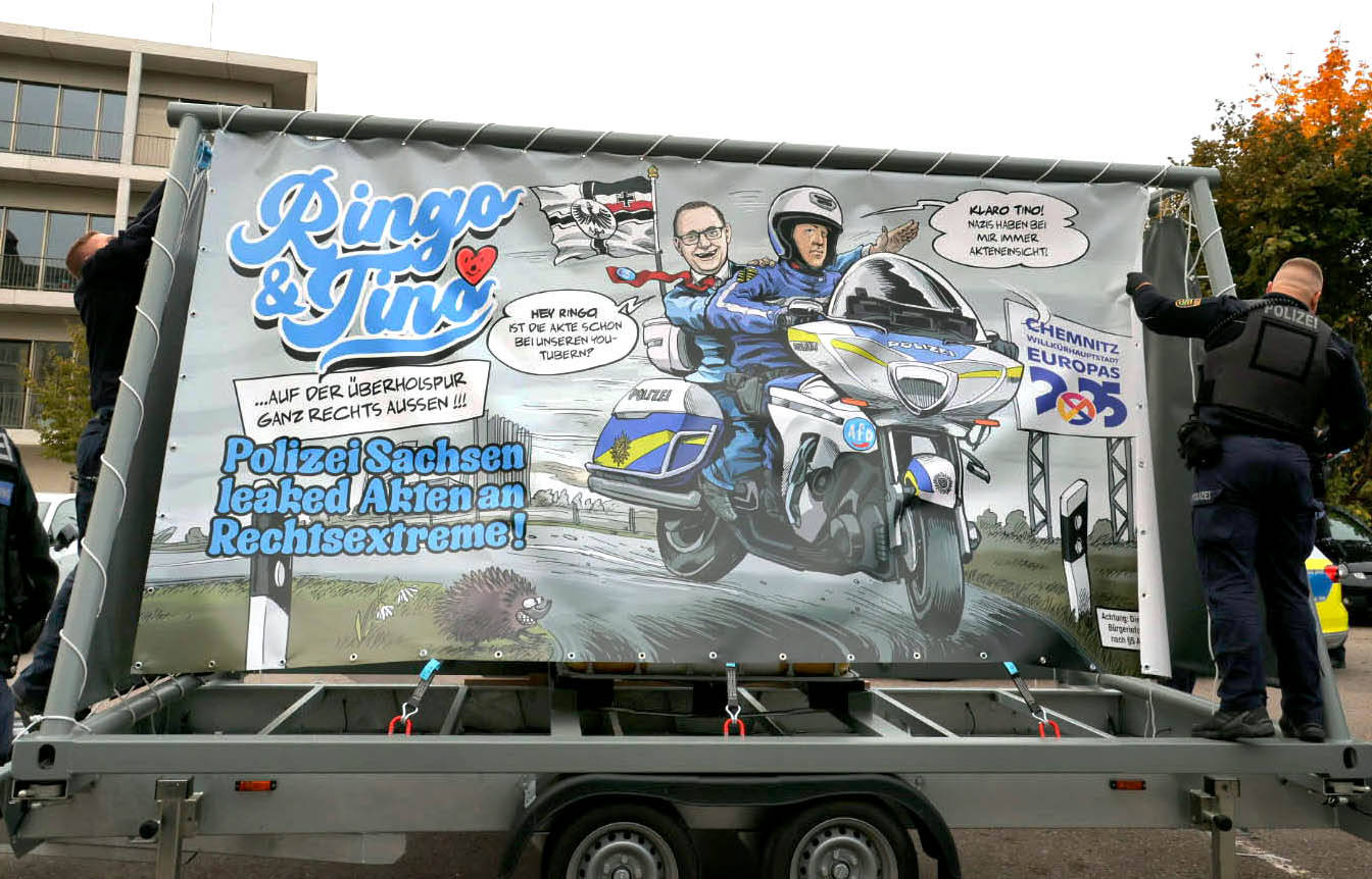 Das Bild zeigt einen Anhänger, auf dem ein großes Banner befestigt ist. Auf dem Banner ist eine Karikatur von zwei Polizisten auf einem Motorrad abgebildet. Der eine Polizist trägt eine Uniform und einen Helm, während der andere einen Anzug und eine Krawatte trägt. Im Hintergrund sind eine Flagge und eine stilisierte Skyline zu sehen. Sprechblasen sind enthalten, die satirische Dialoge zwischen den Figuren enthalten:
