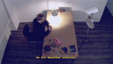 Draufsicht eines trostlosen Verhörraums aus Perspektive einer Überwachungskamera. Sebastian Meineck sitz am Tisch und blickt auf eine Kamera. Auf dem Tisch Utensilien: Lampe, rose Telefon, vertrocknete Pflanze, BND Schreibblock und eine Digitaluhr ist auf 13:12 stehen geblieben.