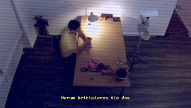 Draufsicht eines trostlosen Verhörraums aus Perspektive einer Überwachungskamera. Marksu Reuter sitzt am Tisch und blickt auf eine Kamera. Auf dem Tisch Utensilien: Lampe, rose Telefon, vertrocknete Pflanze, BND Schreibblock und eine Digitaluhr ist auf 13:12 stehen geblieben.