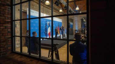 durch eine Fensterfront sind Emmanuel Macron und Friedrich Merz auf einem Podest bei einer Pressekonferenz zu sehen