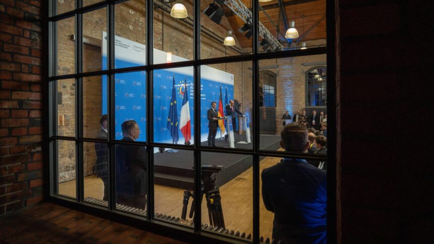 durch eine Fensterfront sind Emmanuel Macron und Friedrich Merz auf einem Podest bei einer Pressekonferenz zu sehen