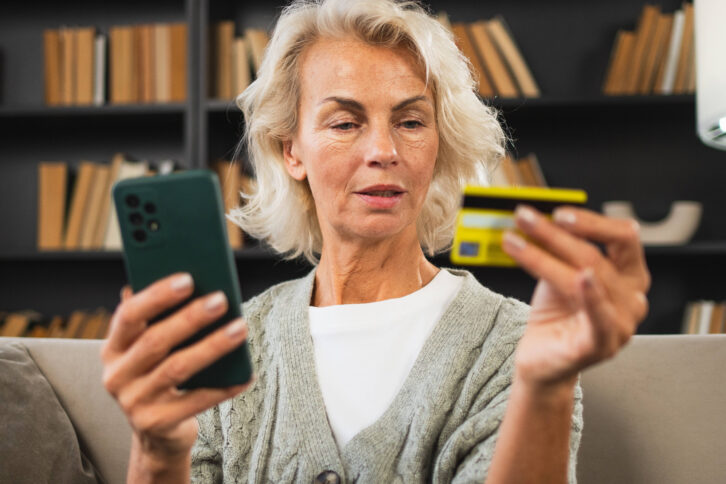 Eine Person mit schulterlangen blonden Haaren hält in der einen Hand eine Kreditkarte und in der anderen ein Smartphone.