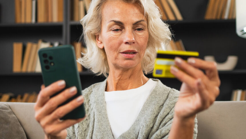 Eine Person mit schulterlangen blonden Haaren hält in der einen Hand eine Kreditkarte und in der anderen ein Smartphone.