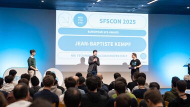 Jean-Baptiste Kempf steht in der Mitte einer Bühne, rechts und links zwei weitere Personen.