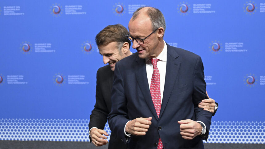 Emmanuel Macron steht hinter Friedrich Merz und fasst ihn am Arm; sie stehen vor einer Fotowand und schauen lächelnd zur Seite