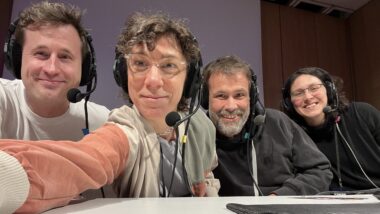 Vier lächelnde Menschen mit Headsets auf dem Kopf, eine Person streckt den Arm nach vorne und nimmt das Selfie auf