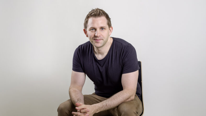 Ein Mann im schwarzen T-Shirt sitzt nach vorne, auf die Knie gebeugt auf einem Stuhl und blickt in die Kamera