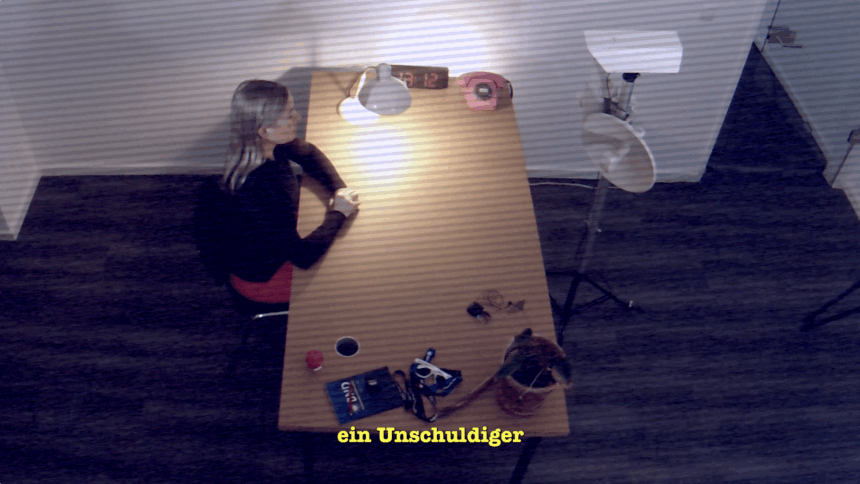 Vogelperspektive aus einer Überwachungskamera in einen tristen Verhörraum: Constanze Kurz sitzen am Tisch mit Blick in eine Kamera.