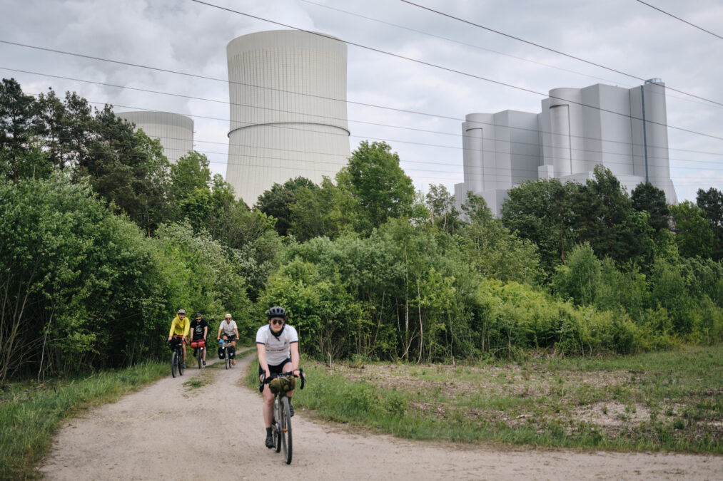 Im Vordergrund Radfahrerinnen vor grünen Bäumen, im Hintergrund graue Kühltürme