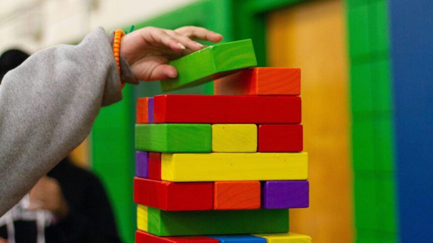 bunte Holzblöcke, die wie beim Spiel Tonga einen Turm bilden; eine Hand legt einen Holzblock auf oben auf den Turm