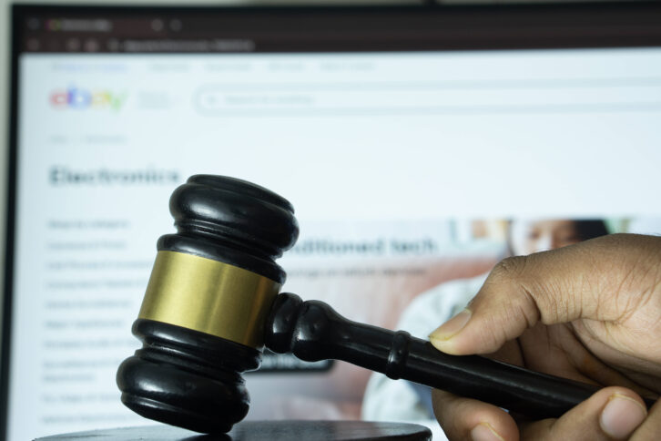 Eine Hand schlägt mit einem Richter*innenhammer auf einen Holzblock. Im Hintergrund ist die Webseite eines Online-Marktplatzes auf einem Latop.