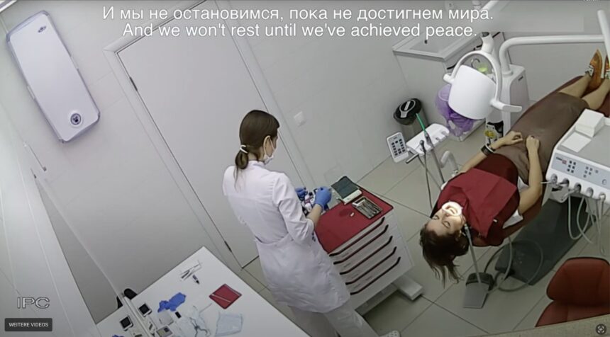 Eine Patientin liegt mit einem Mundöffner auf einer Liege, eine Person in weißem Kittel steht daneben. Screenshot einer Überwachungskamera in einer Zahnarztpraxis in Russland.
