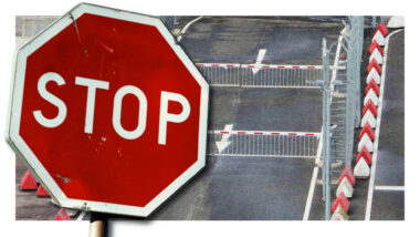 Ein Stoppschild im Vordergrund. Im Hintergrund eine Fahrbahn mit zwei Schranken.