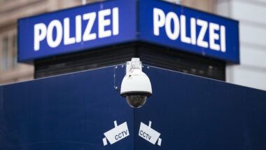 Das Bild zeigt ein blaues Schild mit der Aufschrift "Polizei" in weißer Schrift. Darunter befindet sich eine Überwachungskamera und kleine CCTV-Logos. Im Hintergrund sind Gebäude zu erkennen.