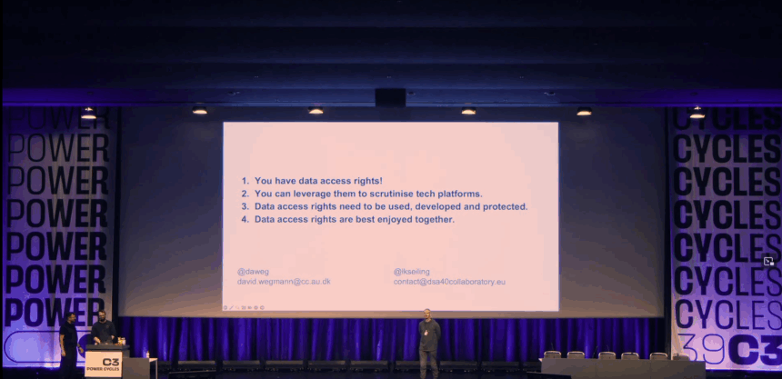 2 Vortragende und 1 Moderierender stehen vor einer großen Beamer-Leinwand