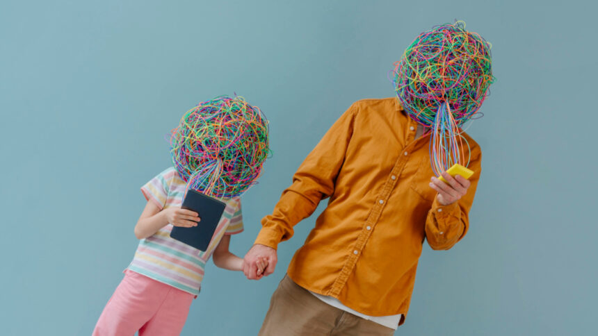 Ein Kind und eine erwachsene Person halten sich an den Händen. Ihre Köpfe sind mit bunten Fäden umwickelt. Sie schauen auf digitale Geräte.