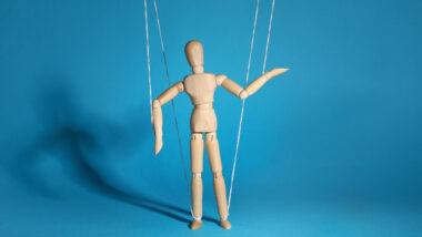 Das Bild zeigt eine Marionette an Fäden vor einem blauen Hintergrund.