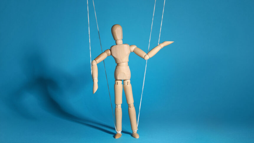 Das Bild zeigt eine Marionette an Fäden vor einem blauen Hintergrund.