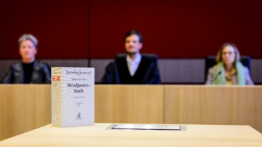 Verschwommenes Foto von zwei Schöff:innen und einem Richter, im Vordergrund ein Strafgesetzbuch