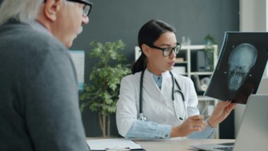 Eine Ärztin weist auf eine Röntgenaufnahme, die einen Schädel zeigt. Ein älterer Mensch hört ihr zu,