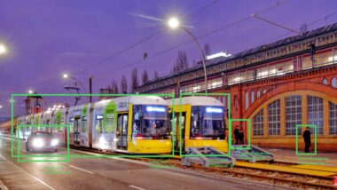 Eine Straße am Abend, in der Mitte zwei gelbe Trams, im Vordergrund Autos, im Hintergrund Fußgänger. Um einige Objekte sind grüne Kästen gezogen, an denen Labels wie "Car" oder "Tram" und "Confidence: 95" stehen