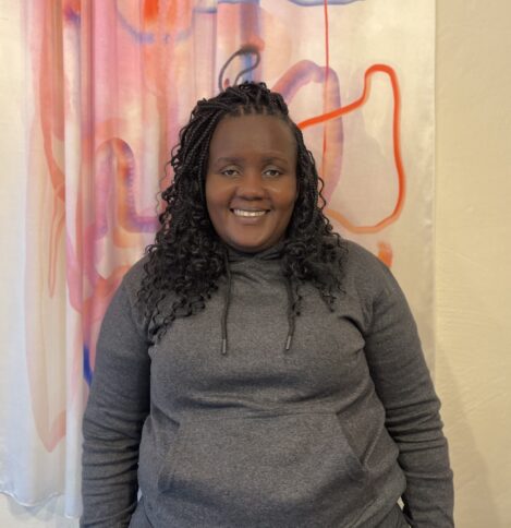 Eine Person mit lockigen Haaren lächelt in die Kamera, im Hintergrund ein abstraktes Bild im lila und rot