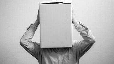 Ein Bild in Grautönen, auf dem eine Person im Hemd zu sehen ist, die sich einen Pappkarton über den Kopf gezogen hat.