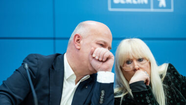 Zwei Berliner Politiker*innen sitzen auf einem Podium und flüstern miteinander Schulter an Schulter hinter vorgehaltenen Händen.