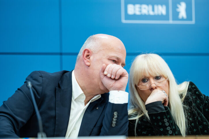 Zwei Berliner Politiker*innen sitzen auf einem Podium und flüstern miteinander Schulter an Schulter hinter vorgehaltenen Händen.