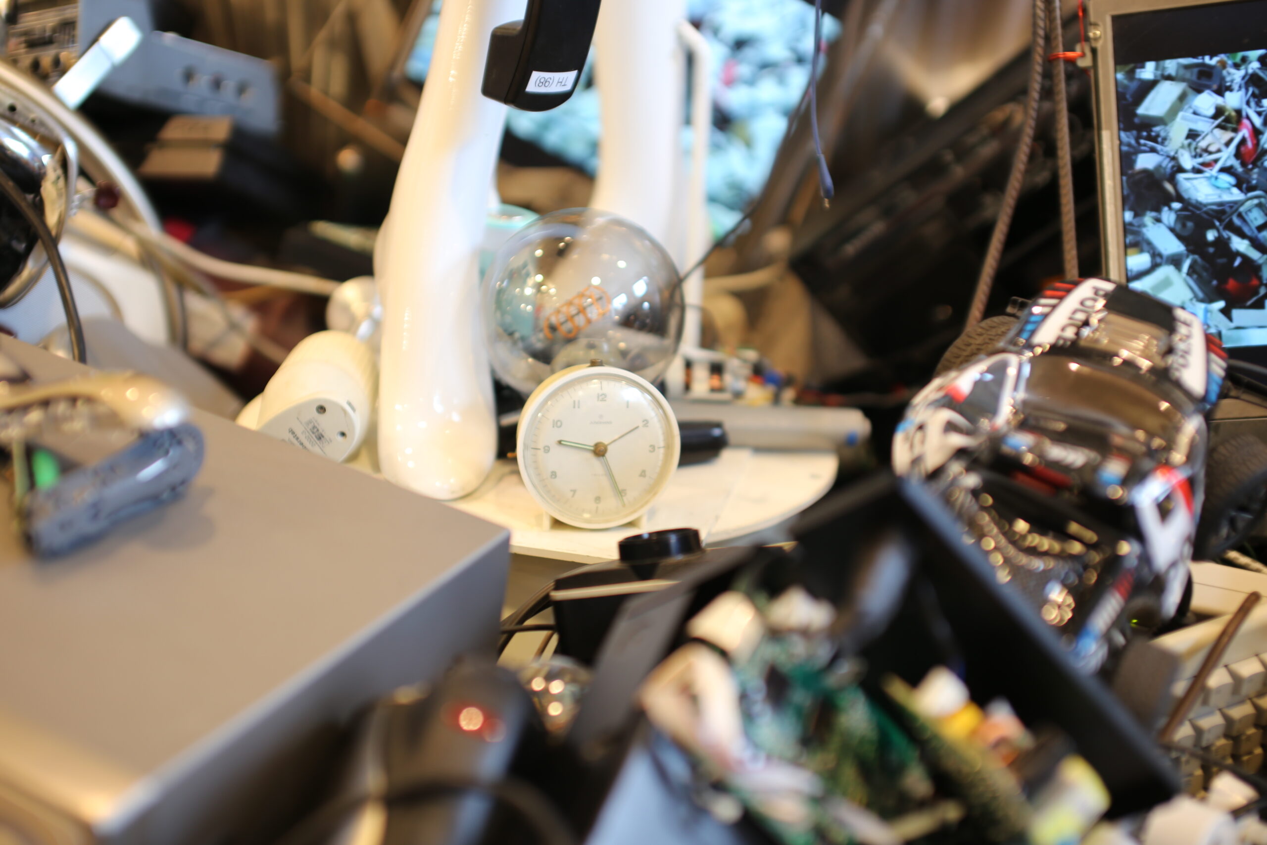 Es sind die Füße der Schaufensterpuppe zu sehen, zwischen denen ein Uhren Wecker steht. Um die Füße herum liegen elektronische Altgeräte.