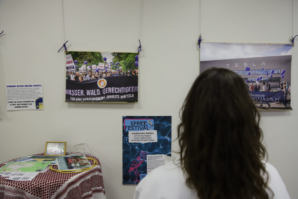 Eine Person mit langen, dunklen, lockigen Haaren steht vor einer Wand mit Plakaten und Fotos, auf denen zum Beispiel demonstrierende Menschen zu sehen sind