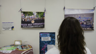 Eine Person mit langen, dunklen, lockigen Haaren steht vor einer Wand mit Plakaten und Fotos, auf denen zum Beispiel demonstrierende Menschen zu sehen sind