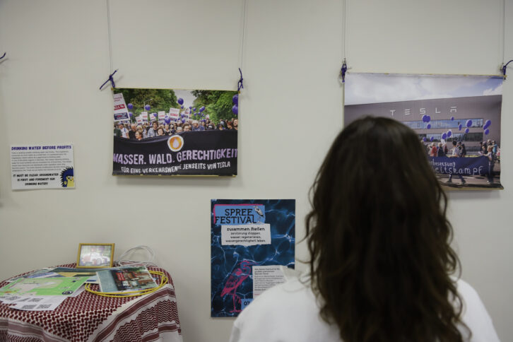 Eine Person mit langen, dunklen, lockigen Haaren steht vor einer Wand mit Plakaten und Fotos, auf denen zum Beispiel demonstrierende Menschen zu sehen sind