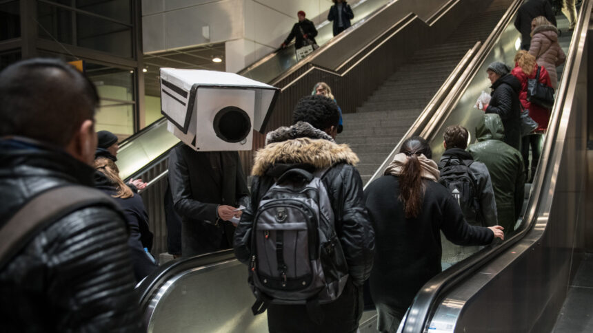 Große Kamera aus Pappe schaut auf Menschen auf einer Rolltreppe.