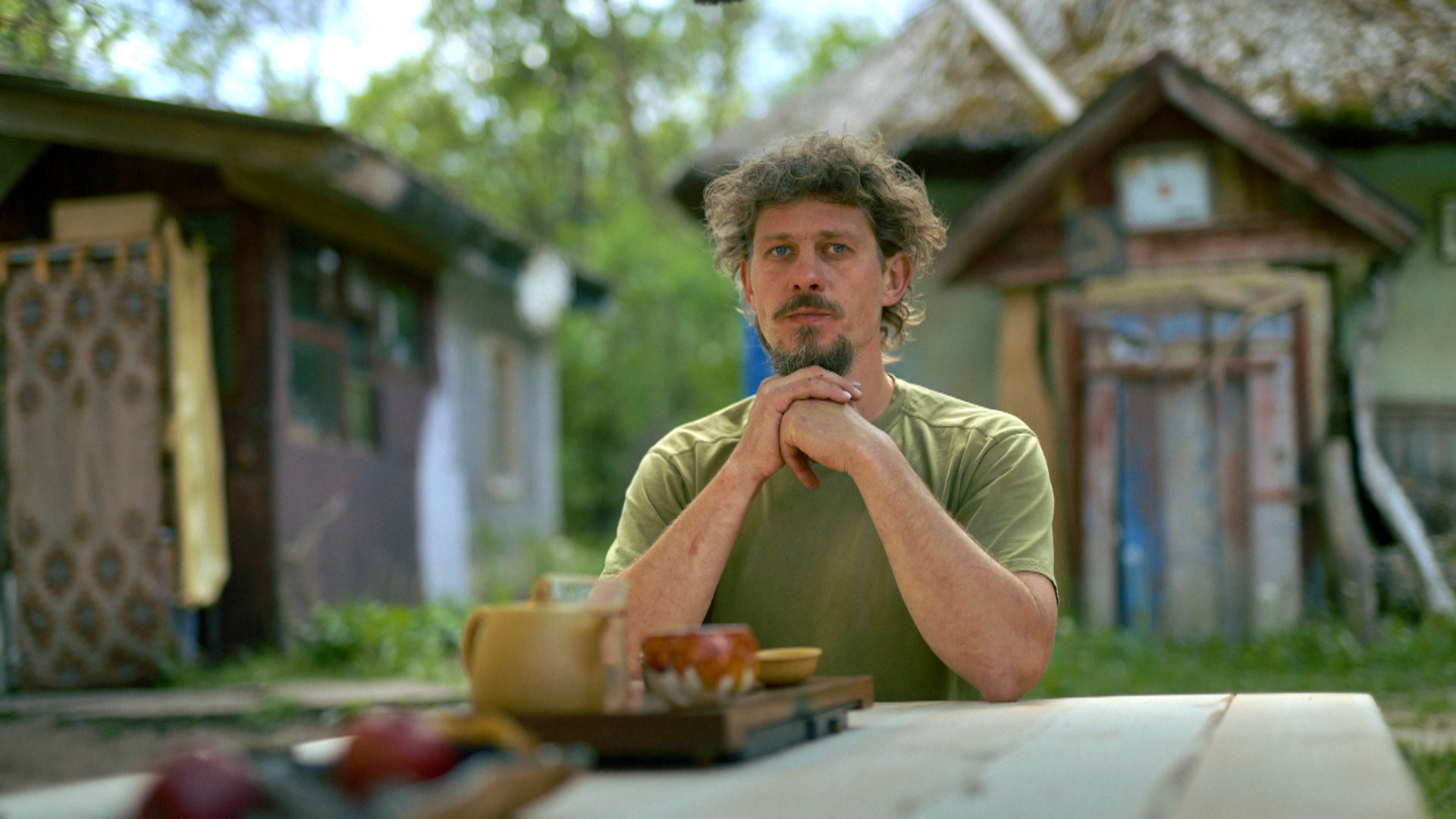 Ein Mann mit Bart sitzt an einem Holztisch auf einem grünen Hof.