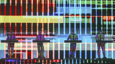 Vier Musiker stehen hinter elektronischen Instrumenten vor einer großen, farbigen, geometrischen Lichtprojektion