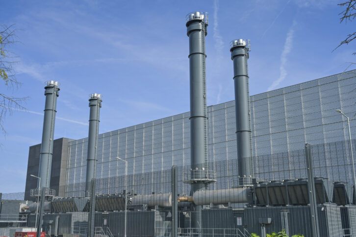 Ein großes graues Industriegebäude mit Schornsteinen und dicken Rohren hinter einem Zaun.