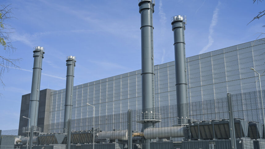 Ein großes graues Industriegebäude mit Schornsteinen und dicken Rohren hinter einem Zaun.