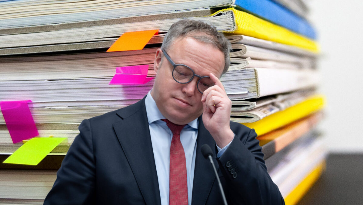 Ein Mann im Anzug steht vor einem Stapel Dokumentenmappen, die mit farbigen Haftnotizen markiert sind.