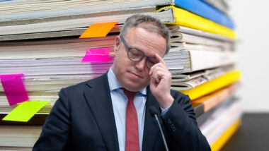 Ein Mann im Anzug steht vor einem Stapel Dokumentenmappen, die mit farbigen Haftnotizen markiert sind.