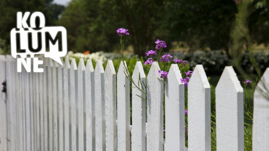 Ein weißer Gartenzaun, zwischen den Latten lugt eine Blume hervor.
