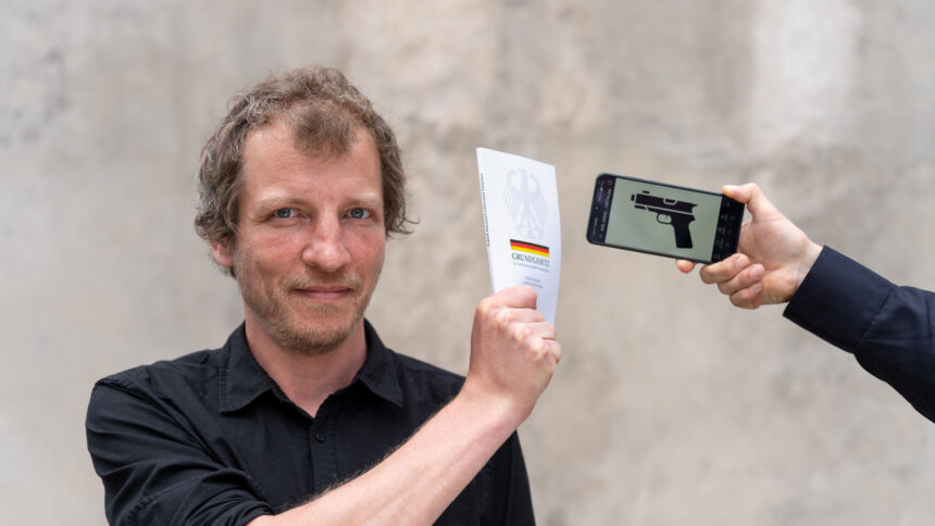 Ein Mann hält das Grundgesetz zwischen sich und ein Mobiltelefon, auf dessen Display eine stilisierte Pistole zu sehen ist.