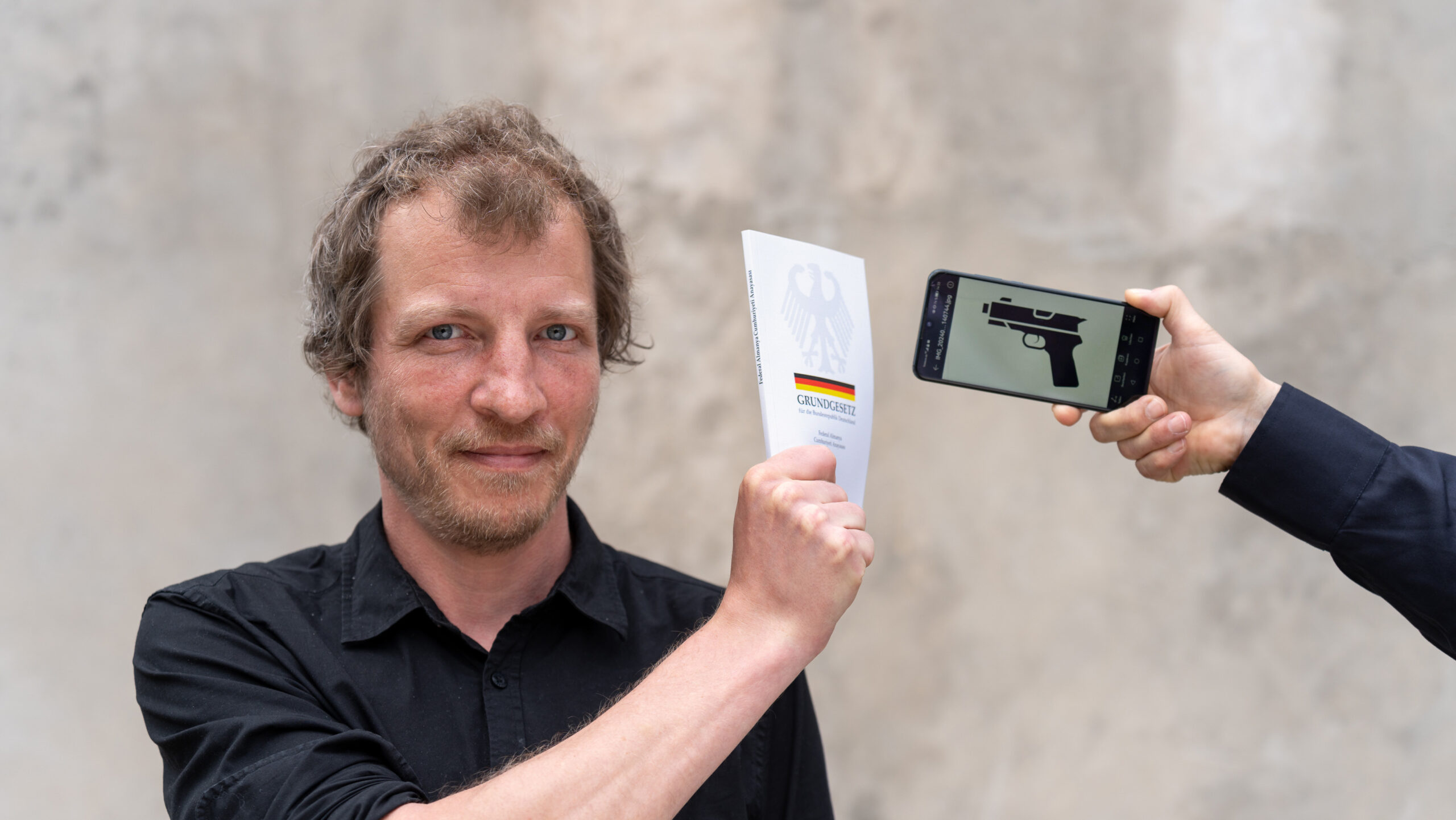 Ein Mann hält das Grundgesetz zwischen sich und ein Mobiltelefon, auf dessen Display eine stilisierte Pistole zu sehen ist.