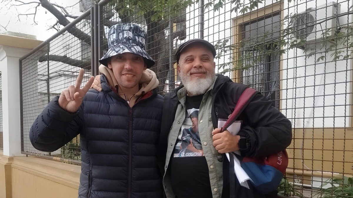 Zwei Personen mit Kopfbedeckungen stehen vor einem Zaun, eine Person macht das Victory-Zeichen mit der Hand